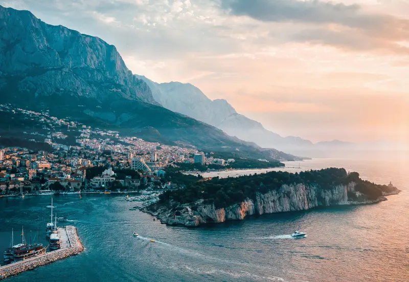 A stunning coastal town nestled between the Adriatic Sea and dramatic mountains, with boats docked in the harbor during a picturesque sunset.

