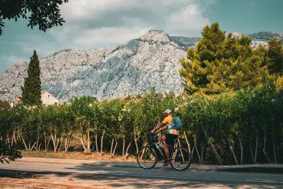 A cyclist riding along a scenic road lined with greenery, with rocky mountains and a clear sky in the background.