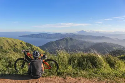 Cyclist sitting on a grassy hilltop with a mountain bike, overlooking rolling green hills, a vast body of water, and distant mountains under a clear blue sky.